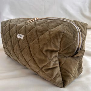 May include: Olive green quilted corduroy cosmetic bag with a gold zipper. The bag has a rectangular shape and a diamond pattern. A small white tag with the text "Elegna" is sewn on the side. The bag is designed for storing makeup and toiletries.