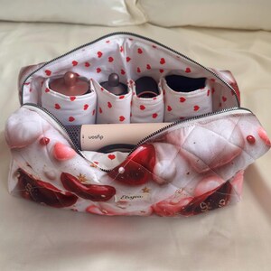 May include: Open cosmetic bag with a quilted exterior featuring a pattern of red hearts and abstract designs. The interior has compartments and a white lining with red hearts. Contains beauty tools and a Dyson hair styling tool.