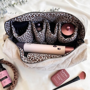 Peut inclure: Trousse de maquillage en velours c&ocirc;tel&eacute; beige avec doublure et compartiments &agrave; imprim&eacute; l&eacute;opard pour les outils de coiffure. Un s&egrave;che-cheveux Dyson est &agrave; l'int&eacute;rieur du sac. Des produits de maquillage et un pinceau sont &agrave; c&ocirc;t&eacute; du sac.