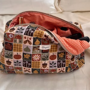 Può includere: Un beauty case con cerniera con un design patchwork con illustrazioni a tema autunnale. L'esterno presenta quadrati con funghi, foglie e zucche. La fodera interna è arancione con pois bianchi. La borsa è aperta, rivelando un rossetto e altri oggetti.