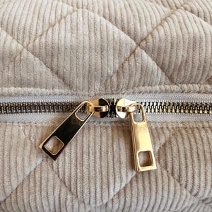 Peut inclure: Gros plan sur un tissu beige matelass&eacute; avec une fermeture &eacute;clair dor&eacute;e. La fermeture &eacute;clair est partiellement ouverte, r&eacute;v&eacute;lant les dents. Le tissu a une texture en velours c&ocirc;tel&eacute; et un motif en losange.