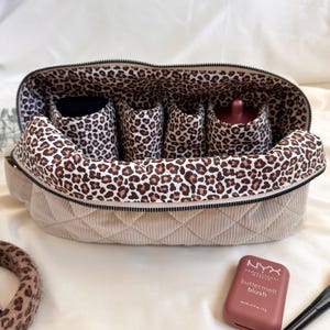 Peut inclure: Trousse de maquillage beige matelass&eacute;e avec int&eacute;rieur et s&eacute;parateurs &agrave; imprim&eacute; l&eacute;opard. Le sac est ouvert, r&eacute;v&eacute;lant plusieurs compartiments et un flacon. Un blush NYX Butter Melt est &agrave; c&ocirc;t&eacute; du sac.