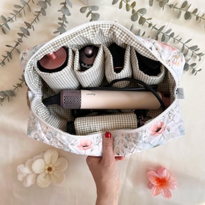 May include: Floral print cosmetic bag with multiple compartments, holding hair styling tools. The bag features a white zipper and a gingham interior. A Dyson hair styler is visible. The bag is decorated with pink flowers and green leaves.