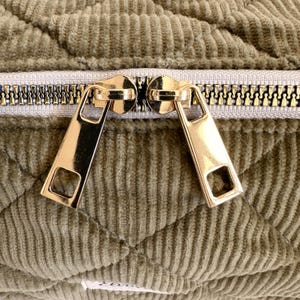 May include: Close-up of a quilted olive green fabric with a gold zipper. The zipper pulls are rectangular with a square cut-out. The fabric has a corduroy texture and a diamond pattern.