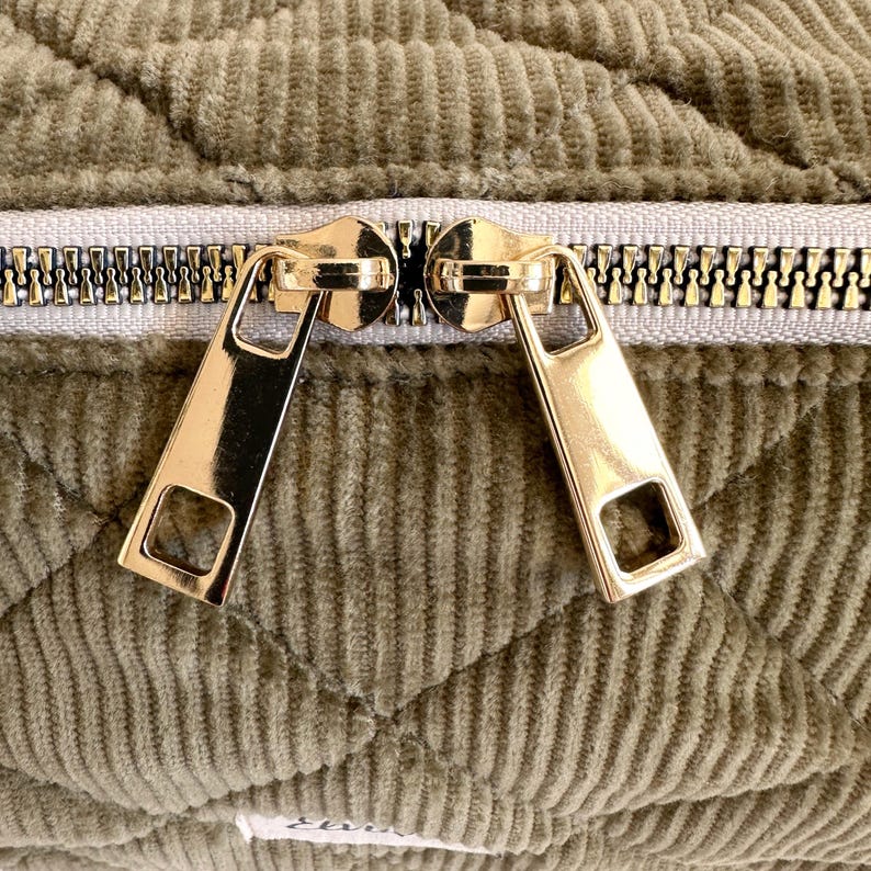 May include: Close-up of a quilted olive green fabric with a gold zipper. The zipper pulls are rectangular with a square cut-out. The fabric has a corduroy texture and a diamond pattern.