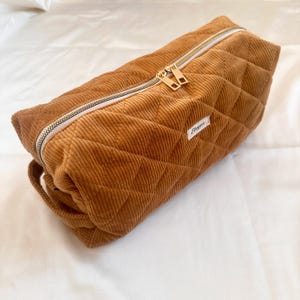 May include: A rectangular, quilted corduroy bag in a warm brown hue. The bag features a gold zipper and a small white label with cursive text. The corduroy has a ribbed texture, and the bag is designed with a handle.