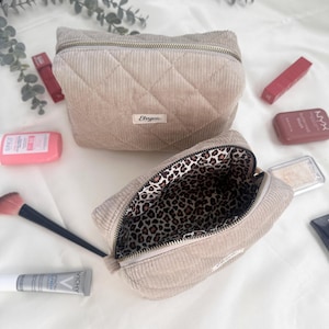 May include: Two beige corduroy cosmetic bags with quilted diamond stitching. One bag is open, revealing a leopard print lining. The bags are surrounded by makeup products, including lipsticks and a makeup brush.