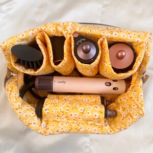 May include: A yellow floral print travel organizer filled with hair styling tools. The organizer contains a hair dryer, brush, and other attachments. The hair dryer is a light pink color with the brand name "Dyson" printed on it.