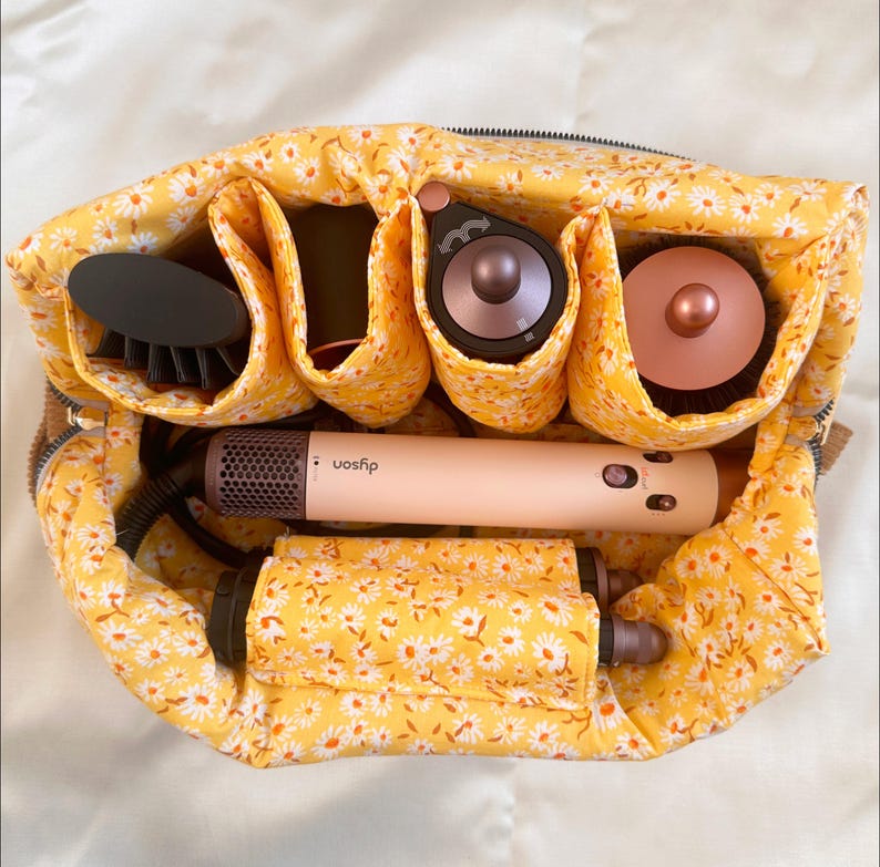 May include: A yellow floral print travel organizer filled with hair styling tools. The organizer contains a hair dryer, brush, and other attachments. The hair dryer is a light pink color with the brand name "Dyson" printed on it.