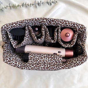 Peut inclure: Un sac de voyage avec un imprim&eacute; l&eacute;opard, dot&eacute; de plusieurs compartiments, contenant un s&egrave;che-cheveux, des brosses et des accessoires. Le sac est ouvert, r&eacute;v&eacute;lant le contenu, sur fond blanc.