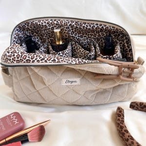 Peut inclure: Une trousse de maquillage en velours c&ocirc;tel&eacute; beige avec un motif matelass&eacute; et un int&eacute;rieur imprim&eacute; l&eacute;opard. Le sac a une fermeture &eacute;clair et plusieurs compartiments. Une pince &agrave; cheveux et des produits de maquillage sont visibles.