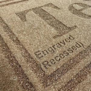 May include: Close-up of a textured, beige rug with the letter "T" and the number "9" in a darker brown hue. The words "Engraved (Recessed)" are also visible, indicating a design feature.