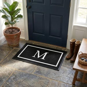 May include: A rectangular black doormat featuring a white border and a large white letter M centered in the middle. The mat has a textured surface and is positioned on a stone floor in front of a dark blue door with a potted plant and boots.