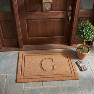 May include: A rectangular tan coir doormat featuring a large embossed letter G in the center, framed by a double-line border. The mat is positioned on a stone patio in front of a dark wood door with a potted green shrub and work gloves.