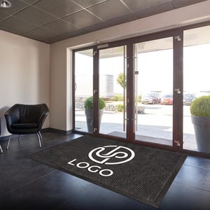 May include: A rectangular dark gray floor mat featuring a white circular logo design with the text "LOGO" printed underneath. The mat is positioned on a dark tiled floor in front of a set of glass double doors leading to an outdoor parking area.