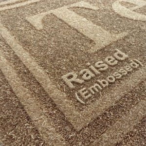May include: Close-up of a textured, brown doormat with the word "Raised (Embossed)" in a lighter shade of brown. The word "TO" is also visible in the upper portion of the mat. The mat has a woven appearance.