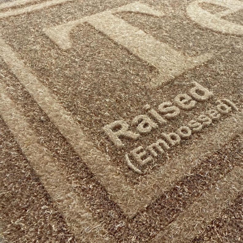 May include: Close-up of a textured, brown doormat with the word "Raised (Embossed)" in a lighter shade of brown. The word "TO" is also visible in the upper portion of the mat. The mat has a woven appearance.