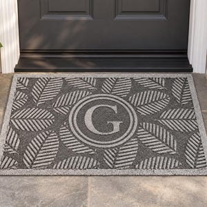 May include: A rectangular doormat featuring a dark gray background with a repeating pattern of light gray leaves. In the center, a circular emblem encloses the capital letter G in a light gray font, surrounded by a light gray border.