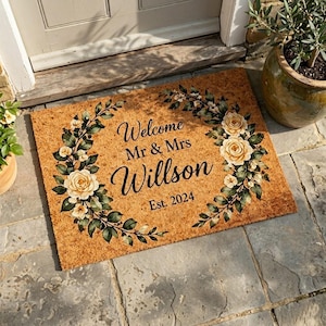 May include: A rectangular coir doormat with a floral wreath design. The mat features the text "Welcome Mr & Mrs Willson Est. 2024" in black script. The wreath includes cream-colored roses and green leaves.