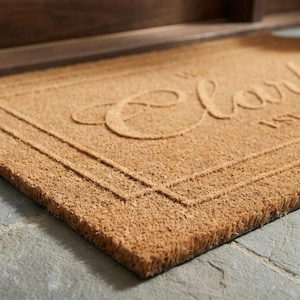 May include: A rectangular doormat made of natural coir fibers. The mat is a light brown color with a raised border and the name "Clark" in a cursive font. The mat is placed on a gray stone surface.
