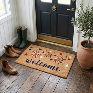 May include: A rectangular coir doormat with the word "welcome" in navy blue script, decorated with red, white, and blue fireworks and stars. Brown leather boots and green rain boots are placed on the wooden floor near the door.