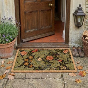 May include: A rectangular doormat featuring a dark background with a dense pattern of large green leaves and orange flowers. The mat has a natural tan fiber border and is positioned on a stone floor in front of a wooden door with a brass knob.