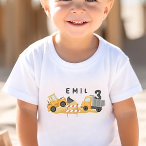 May include: A white short-sleeved t-shirt featuring a graphic print of a yellow construction excavator and a dump truck on a dirt pile. The text "EMIL" is printed above the vehicles, and the number "3" is displayed on the side of the truck.