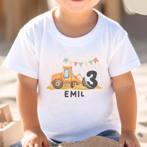 K&ouml;nnte beinhalten: Ein wei&szlig;es T-Shirt mit kurzen &Auml;rmeln, das eine Grafik eines orangefarbenen Baggers, eine gro&szlig;e schwarze Zahl 3 und bunte Wimpel zeigt. Der Name EMIL ist unter der Grafik in Schwarz gedruckt. Ein rundes beiges Etikett zeigt den Text f&uuml;r kleine Bauarbeiter.