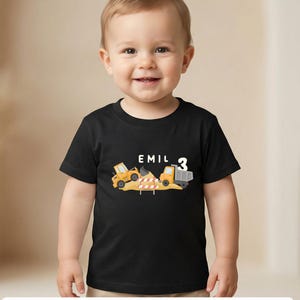May include: A black short-sleeved t-shirt featuring a graphic of a yellow construction excavator and a dump truck on a pile of dirt. The text EMIL is printed in white above the vehicles, with the number 3 displayed in white to the right side.