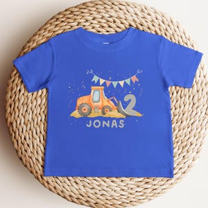 K&ouml;nnte beinhalten: Ein k&ouml;nigsblaues T-Shirt mit kurzen &Auml;rmeln, das eine Grafik eines orangefarbenen Baufahrzeugs, eine gro&szlig;e graue Zahl zwei und bunte Wimpel zeigt. Der Name JONAS ist in Grau unter dem Fahrzeug gedruckt. Der Text ROYAL - BABY SHIRT ist sichtbar.