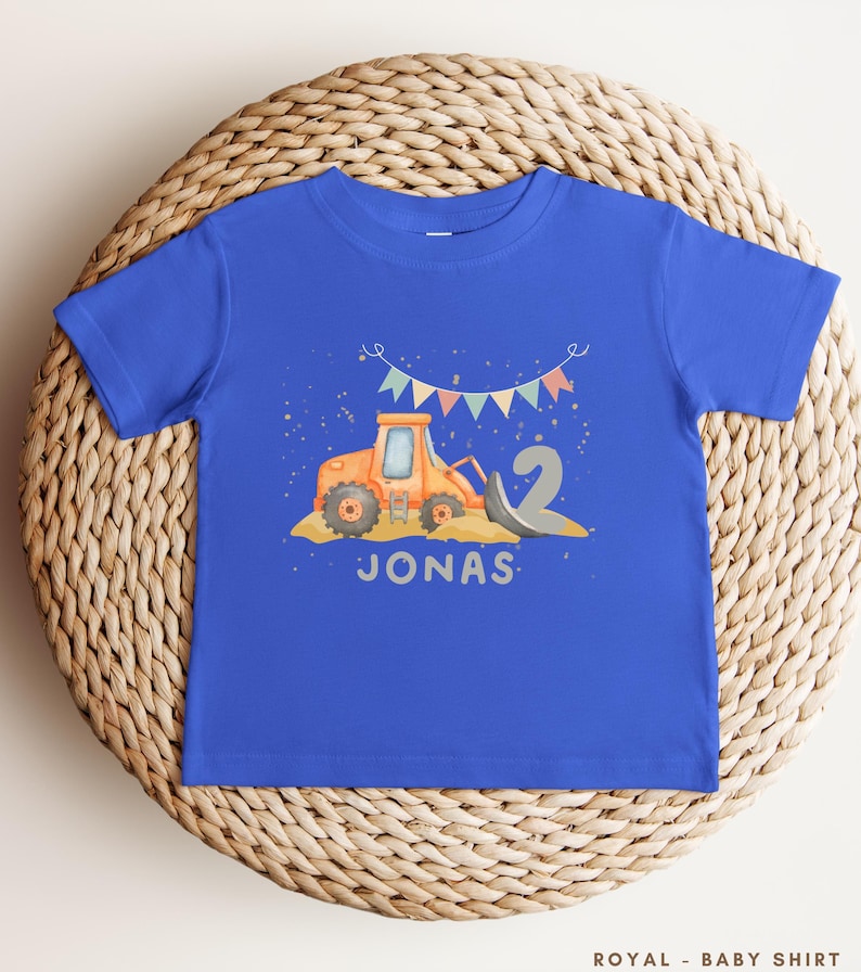 K&ouml;nnte beinhalten: Ein k&ouml;nigsblaues T-Shirt mit kurzen &Auml;rmeln, das eine Grafik eines orangefarbenen Baufahrzeugs, eine gro&szlig;e graue Zahl zwei und bunte Wimpel zeigt. Der Name JONAS ist in Grau unter dem Fahrzeug gedruckt. Der Text ROYAL - BABY SHIRT ist sichtbar.