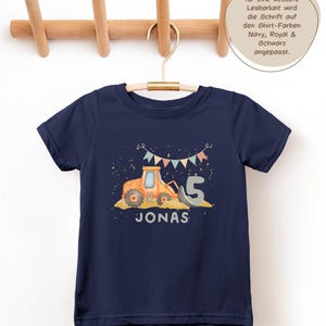 K&ouml;nnte beinhalten: Ein marineblaues Kurzarm-T-Shirt mit einer Grafik eines orangefarbenen Baustellentraktors, einer gro&szlig;en grauen Zahl 5 und dem Namen JONAS in wei&szlig;er Schrift. &Uuml;ber dem Traktor befindet sich eine Girlande aus bunten Wimpeln.