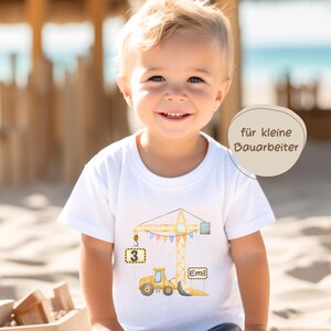 May include: A white short-sleeved t-shirt featuring a graphic of a yellow construction crane and a yellow bulldozer. The crane hook holds a square sign with the number 3, and a rectangular sign displays the name Emil. Text reads f&uuml;r kleine Bauarbeiter.