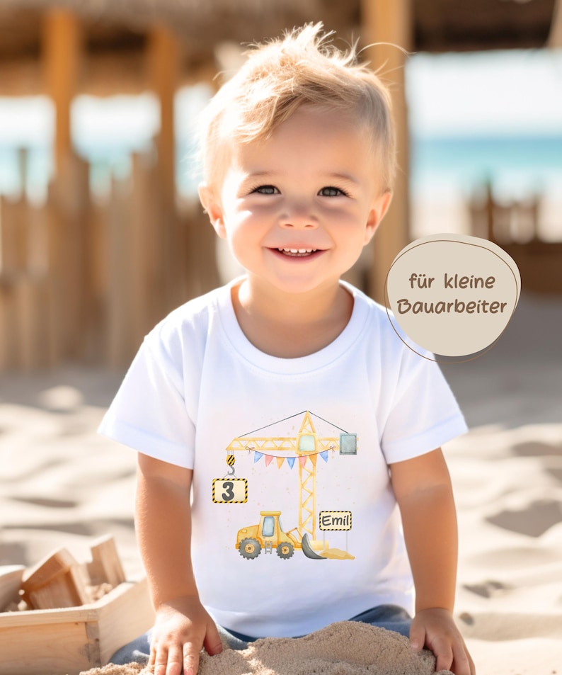 May include: A white short-sleeved t-shirt featuring a graphic of a yellow construction crane and a yellow bulldozer. The crane hook holds a square sign with the number 3, and a rectangular sign displays the name Emil. Text reads f&uuml;r kleine Bauarbeiter.