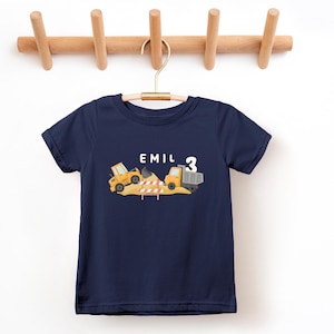 May include: A navy blue short-sleeved t-shirt featuring a graphic of yellow construction vehicles, a pile of dirt, and a red and white striped barrier. The text "EMIL" and the number "3" are printed in white above the construction scene.