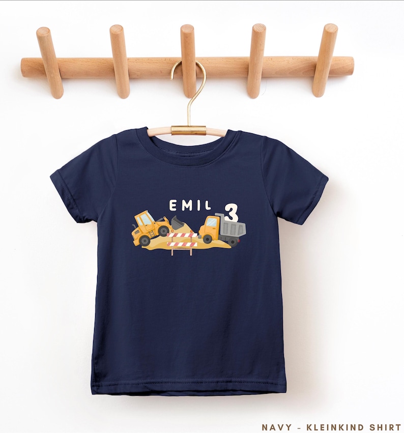 May include: A navy blue short-sleeved t-shirt featuring a graphic of yellow construction vehicles, a pile of dirt, and a red and white striped barrier. The text "EMIL" and the number "3" are printed in white above the construction scene.