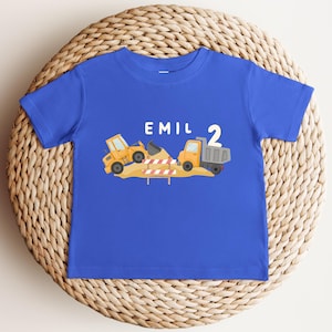 May include: A royal blue short-sleeve t-shirt featuring a graphic of yellow construction vehicles, a pile of dirt, and a red and white striped barrier. The text "EMIL" and the number "2" are printed in white above the construction scene.