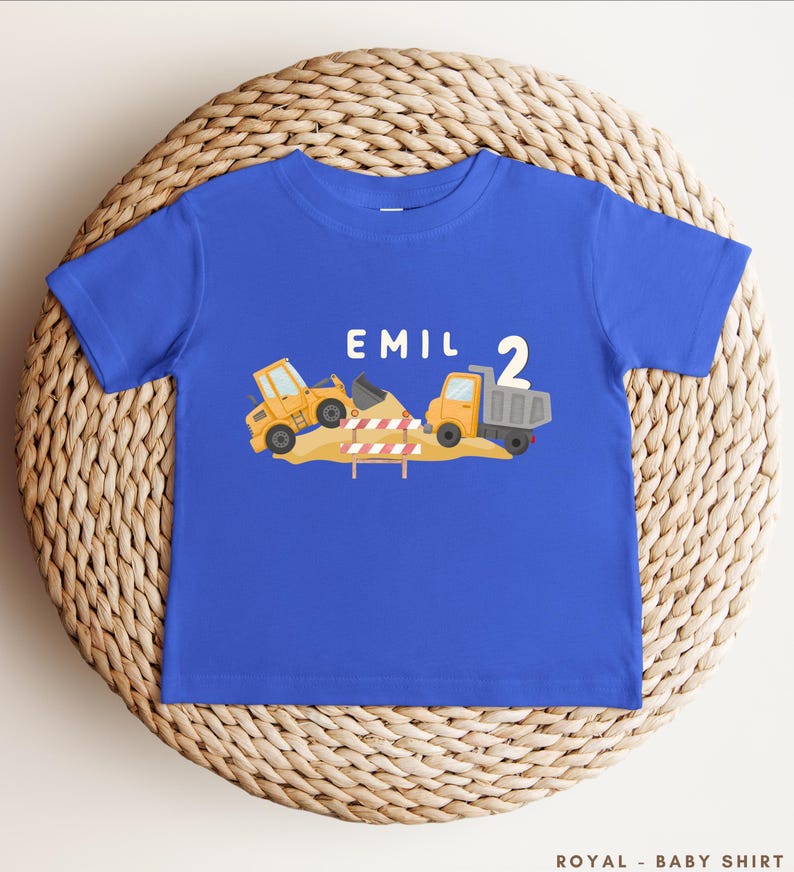 May include: A royal blue short-sleeve t-shirt featuring a graphic of yellow construction vehicles, a pile of dirt, and a red and white striped barrier. The text "EMIL" and the number "2" are printed in white above the construction scene.