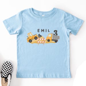 May include: A light blue short-sleeve t-shirt featuring a graphic of a yellow construction loader and a dump truck on a pile of dirt with a red and white striped barrier. The text EMIL and the number 3 are printed in black above the construction vehicles.