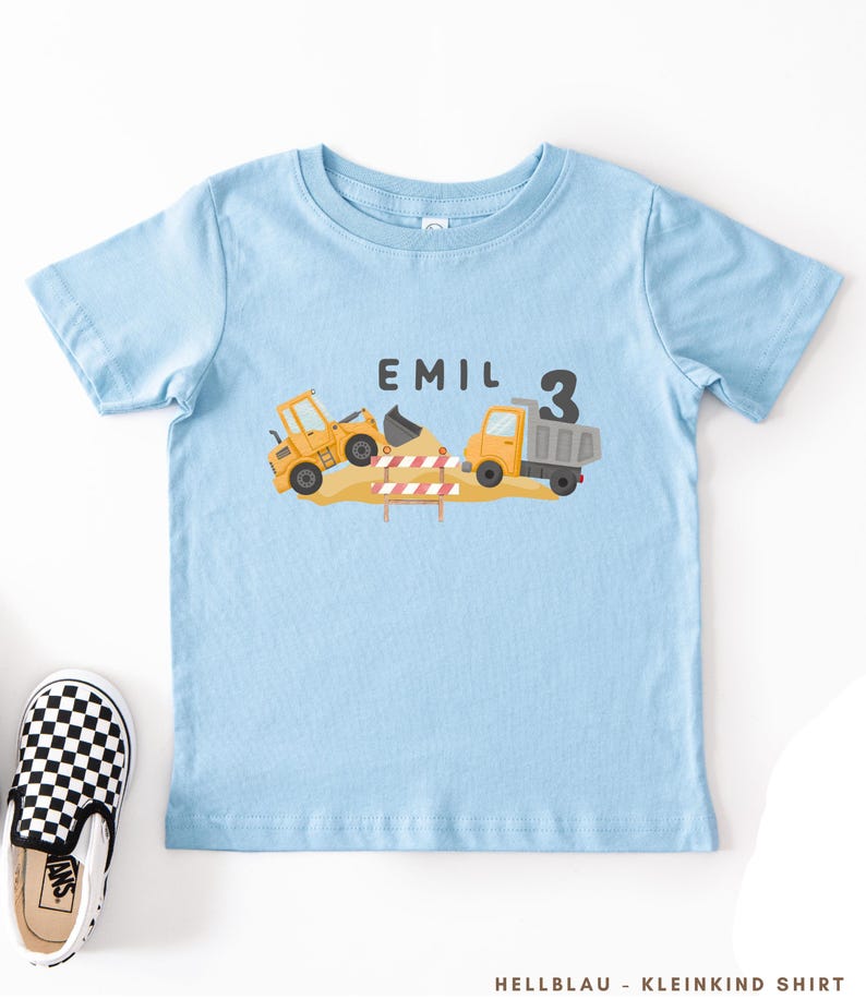 May include: A light blue short-sleeve t-shirt featuring a graphic of a yellow construction loader and a dump truck on a pile of dirt with a red and white striped barrier. The text EMIL and the number 3 are printed in black above the construction vehicles.