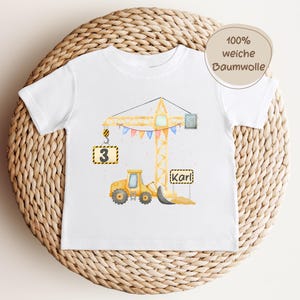 May include: A white short-sleeve t-shirt featuring a watercolor illustration of a yellow construction crane and a yellow bulldozer. The design includes a sign with the number 3, a sign with the name Karl, and colorful bunting flags.