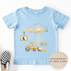 May include: A light blue short-sleeve t-shirt featuring a yellow construction crane graphic with a hanging sign displaying the number 4 and a second sign with the name Emil, decorated with colorful bunting flags and a yellow bulldozer.