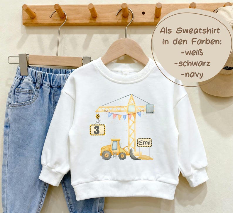 May include: A white long-sleeve sweatshirt featuring a watercolor illustration of a yellow construction crane and a bulldozer. The crane holds a sign with the number 3, and a sign with the name Emil is near the bulldozer. Text reads: Als Sweatshirt in den Farben: -wei&szlig; -schwarz -navy.