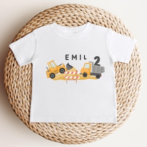 May include: A white short-sleeve t-shirt featuring a graphic of a yellow construction excavator and a dump truck on a dirt pile with a red and white striped barrier. The text EMIL and the number 2 are printed in black above the construction vehicles.