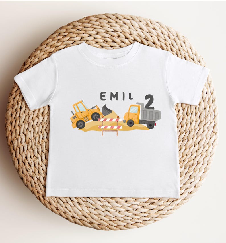May include: A white short-sleeve t-shirt featuring a graphic of a yellow construction excavator and a dump truck on a dirt pile with a red and white striped barrier. The text EMIL and the number 2 are printed in black above the construction vehicles.