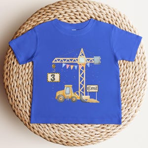 May include: A royal blue short-sleeve t-shirt featuring a graphic illustration of a yellow construction crane and a yellow excavator. The crane holds a sign with the number 3, and a separate sign displays the name Emil. Colorful bunting hangs from the crane.