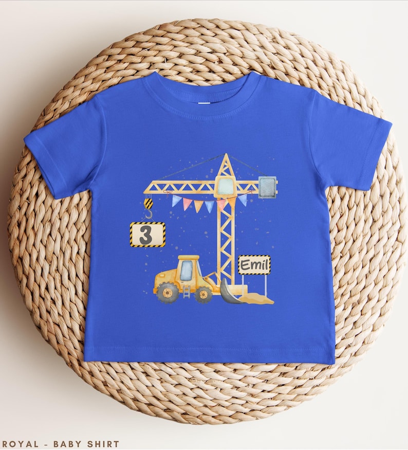 May include: A royal blue short-sleeve t-shirt featuring a graphic illustration of a yellow construction crane and a yellow excavator. The crane holds a sign with the number 3, and a separate sign displays the name Emil. Colorful bunting hangs from the crane.
