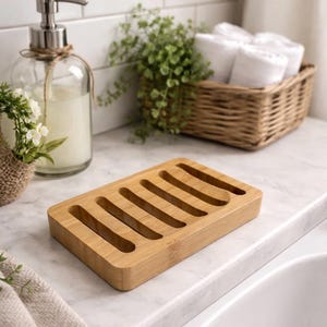 May include: A rectangular bamboo soap dish with six parallel slots. The light brown dish sits on a white countertop, near a glass soap dispenser and a basket of rolled white towels. The background includes white tile and greenery.