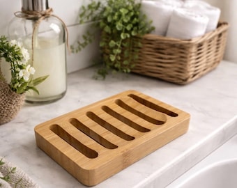 May include: A rectangular bamboo soap dish with six parallel slots. The light brown dish sits on a white countertop, near a glass soap dispenser and a basket of rolled white towels. The background includes white tile and greenery.