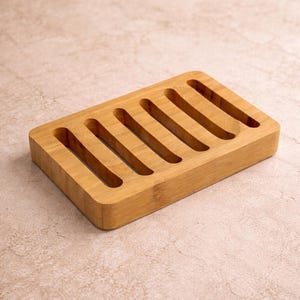 May include: A rectangular bamboo soap dish with seven parallel slots. The dish is made of light brown bamboo and has a simple, minimalist design. The slots allow for drainage, extending the life of the soap. The dish is suitable for the bathroom or kitchen.